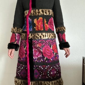 May include: A long black coat with floral and leopard print accents. The coat features a pink velvet trim, black fur cuffs, and a decorative floral pattern in shades of pink, orange, and purple. The coat has a unique, eye-catching design.