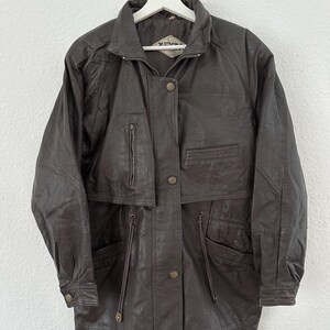 Puede incluir: Chaqueta de cuero marr&oacute;n oscuro con cuello, cremallera y cierre de botones. La chaqueta tiene m&uacute;ltiples bolsillos, incluidos dos bolsillos delanteros y un bolsillo en el pecho. Cuelga de una percha blanca sobre un fondo blanco.