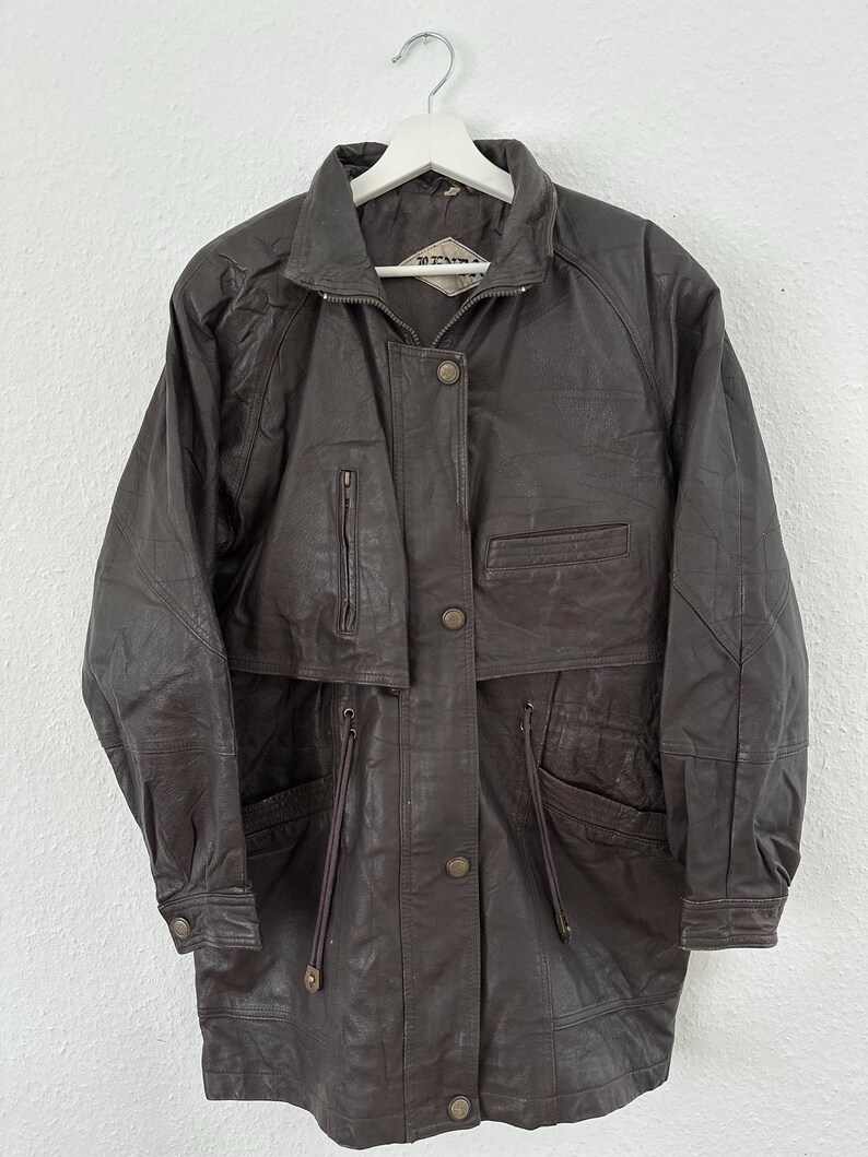 Puede incluir: Chaqueta de cuero marr&oacute;n oscuro con cuello, cremallera y cierre de botones. La chaqueta tiene m&uacute;ltiples bolsillos, incluidos dos bolsillos delanteros y un bolsillo en el pecho. Cuelga de una percha blanca sobre un fondo blanco.