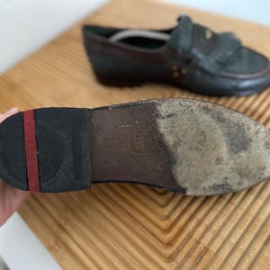Puede incluir: Primer plano de la suela de un mocas&iacute;n de cuero desgastado, sostenido por una mano. La suela es negra con una raya roja y muestra signos de desgaste. Otro mocas&iacute;n est&aacute; en el fondo, con una combinaci&oacute;n de colores azul oscuro y marr&oacute;n.