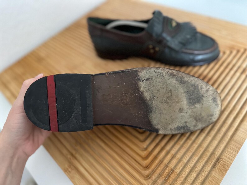 Puede incluir: Primer plano de la suela de un mocas&iacute;n de cuero desgastado, sostenido por una mano. La suela es negra con una raya roja y muestra signos de desgaste. Otro mocas&iacute;n est&aacute; en el fondo, con una combinaci&oacute;n de colores azul oscuro y marr&oacute;n.
