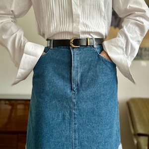 May include: A black leather belt with a gold buckle is worn over a denim skirt. The skirt is a medium blue colour. A white button-down shirt with vertical pleats is worn on top.