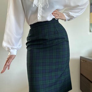 Op de afbeelding: Een witte blouse met een kanten kraag en lange mouwen, gecombineerd met een groen en marineblauwe geruite potloodrok. De rok is getailleerd en komt tot de knieën. De outfit is een klassieke, vintage stijl.