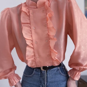 May include: A peach-coloured blouse with a high collar and ruffled trim down the front. The long sleeves have a slight puff at the shoulders and end in ruffled cuffs. The blouse is buttoned down the front and paired with blue jeans and a black belt.