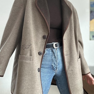 May include: A tan wool coat with a brown trim and buttons. The coat has two front pockets and a leaf design embroidered on the front. The coat is worn over a brown turtleneck and blue jeans.
