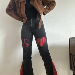 Könnte beinhalten: Braune Lederjacke und schwarze Jeans mit Peace-Zeichen- und Herz-Patches. Die Jeans hat ausgestellte Hosenbeine mit rot-orangefarbenen Stoffeinsätzen. Die Jacke ist im klassischen Biker-Stil gehalten.