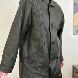 May include: Dark green leather jacket with a button-up front, collared neckline, and two front pockets. The jacket has vertical stitched detailing on the upper chest and sleeves. The jacket is worn over a brown turtleneck.