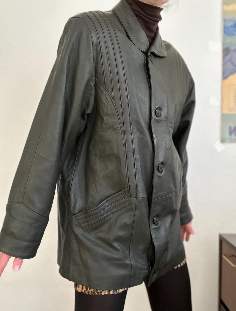 May include: Dark green leather jacket with a button-up front, collared neckline, and two front pockets. The jacket has vertical stitched detailing on the upper chest and sleeves. The jacket is worn over a brown turtleneck.