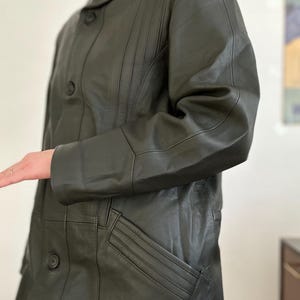 May include: Dark olive green leather coat with a button-up front, a notched collar, and a pocket. The coat has vertical stitching details on the front and sleeves. The coat is a mid-length style.