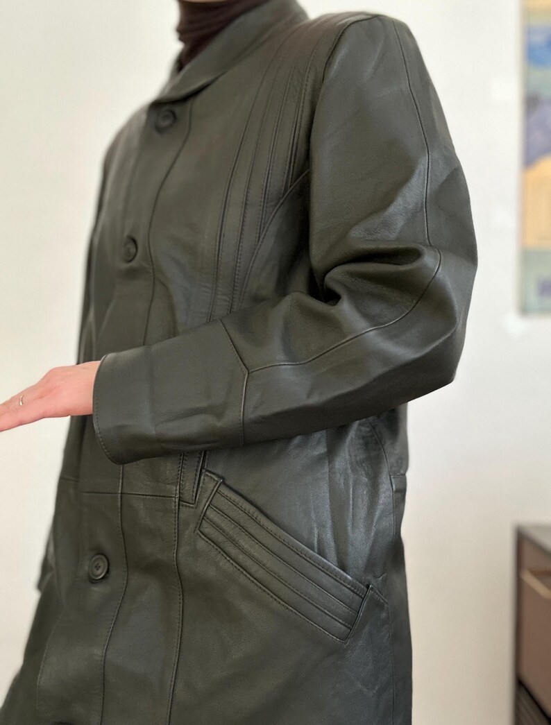May include: Dark olive green leather coat with a button-up front, a notched collar, and a pocket. The coat has vertical stitching details on the front and sleeves. The coat is a mid-length style.