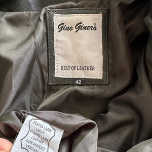 May include: Close-up of the inside of a dark green leather jacket. The label reads "Gino Ginero BEST OF LEATHER" with a size of 42. A second tag indicates "GENUINE LAMB LEATHER".