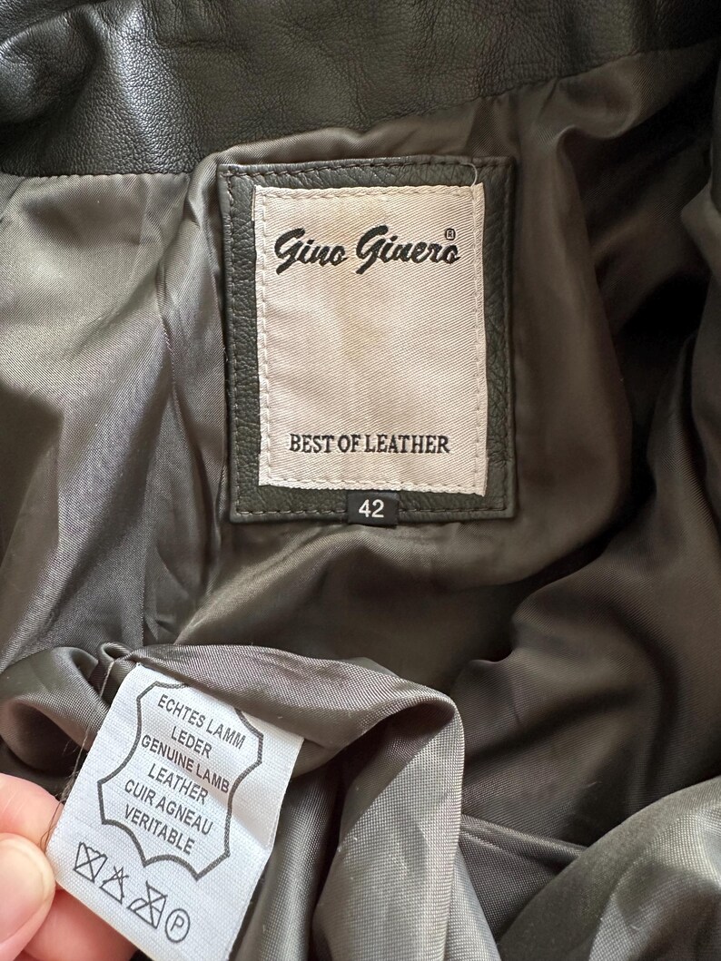 May include: Close-up of the inside of a dark green leather jacket. The label reads "Gino Ginero BEST OF LEATHER" with a size of 42. A second tag indicates "GENUINE LAMB LEATHER".