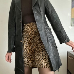 May include: A black leather coat with white stitching and a notched collar. The coat is buttoned and has pockets. The outfit includes a brown turtleneck and a leopard print skirt. The coat is knee-length.