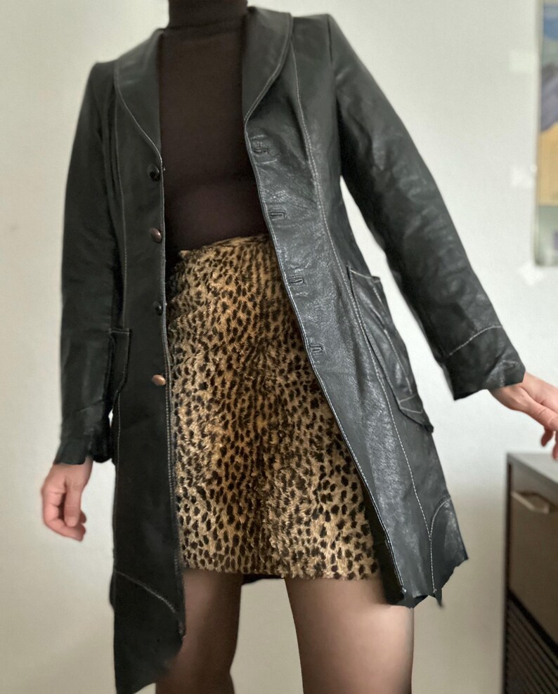 May include: A black leather coat with white stitching and a notched collar. The coat is buttoned and has pockets. The outfit includes a brown turtleneck and a leopard print skirt. The coat is knee-length.