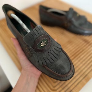 Puede incluir: Un par de mocasines de cuero gris y marr&oacute;n con flecos decorativos y un peque&ntilde;o emblema. Los zapatos tienen un dise&ntilde;o cl&aacute;sico con punta redonda y tac&oacute;n bajo. Un zapato est&aacute; enfocado, el otro est&aacute; ligeramente borroso en el fondo.