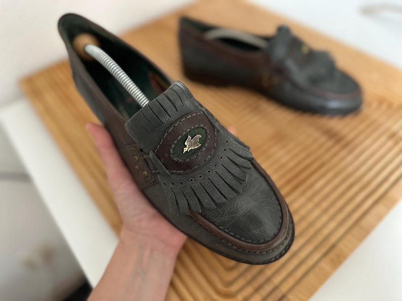 Puede incluir: Un par de mocasines de cuero gris y marr&oacute;n con flecos decorativos y un peque&ntilde;o emblema. Los zapatos tienen un dise&ntilde;o cl&aacute;sico con punta redonda y tac&oacute;n bajo. Un zapato est&aacute; enfocado, el otro est&aacute; ligeramente borroso en el fondo.