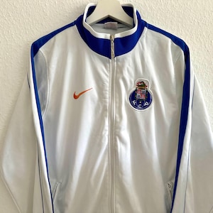 May include: White zip-up track jacket with blue trim and a blue collar. Includes an orange Nike logo and a team emblem. The jacket has a full-length zip and two front pockets.
