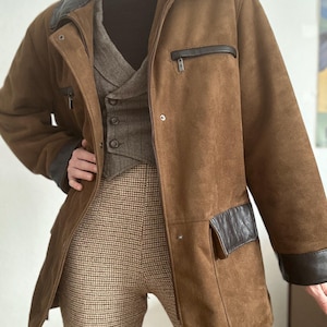 Può includere: Giacca in camoscio marrone con dettagli in pelle nera su colletto, tasche e polsini. La giacca ha una chiusura con zip e una vestibilità su misura. Sotto si vedono un gilet grigio e pantaloni fantasia.