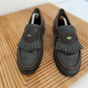 Puede incluir: Un par de mocasines de cuero gris oscuro con ribetes marrones y flecos. Cada zapato presenta un emblema decorativo y est&aacute; colocado sobre una superficie de madera.