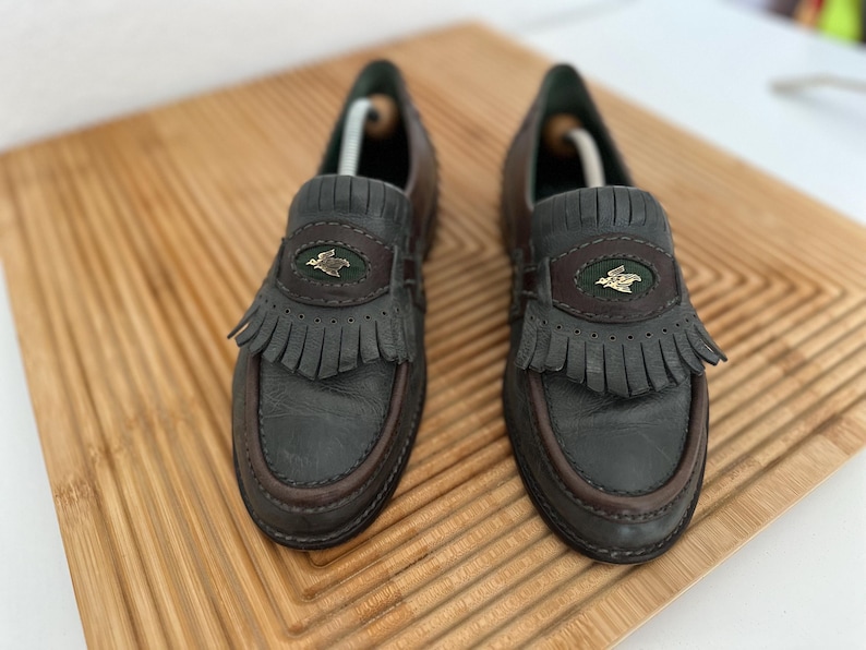 Puede incluir: Un par de mocasines de cuero gris oscuro con ribetes marrones y flecos. Cada zapato presenta un emblema decorativo y est&aacute; colocado sobre una superficie de madera.