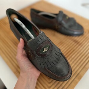 Puede incluir: Un par de mocasines de cuero gris oscuro con flecos decorativos y un peque&ntilde;o emblema. Los zapatos tienen un ribete marr&oacute;n y est&aacute;n colocados sobre una superficie de madera. Un zapato tiene un horma.