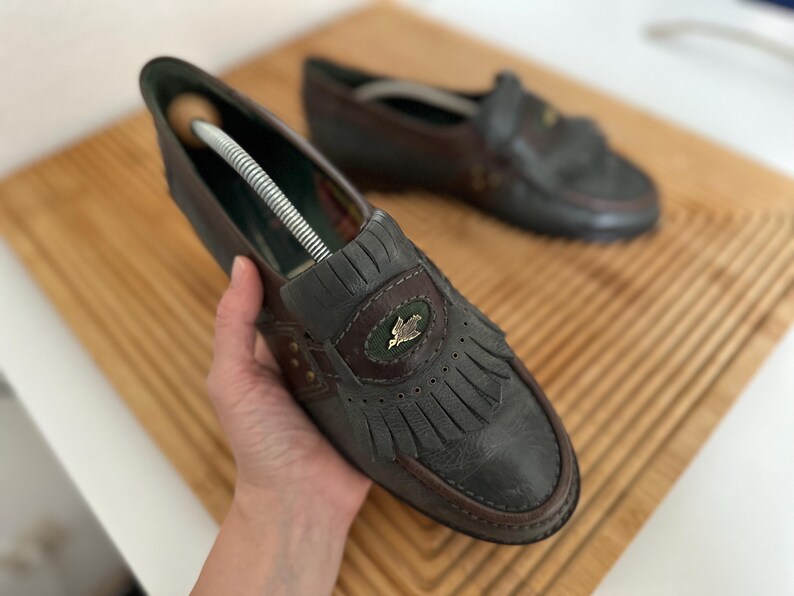 Puede incluir: Un par de mocasines de cuero gris oscuro con flecos decorativos y un peque&ntilde;o emblema. Los zapatos tienen un ribete marr&oacute;n y est&aacute;n colocados sobre una superficie de madera. Un zapato tiene un horma.