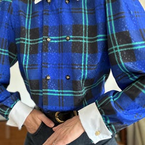 May include: A long-sleeved blouse featuring a blue, black, and teal plaid pattern with a subtle textured finish. The garment includes a crisp white pointed collar, white cuffs with gold-toned buttons, and a double-breasted front with black buttons.