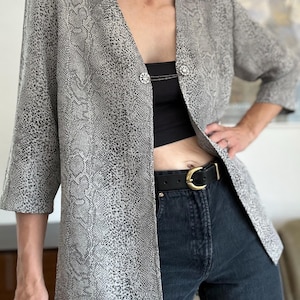 May include: A grey and black snakeskin print blazer featuring three-quarter length sleeves, a V-neckline, and a silver-toned metal clasp closure with a delicate chain detail connecting the two sides of the front opening.