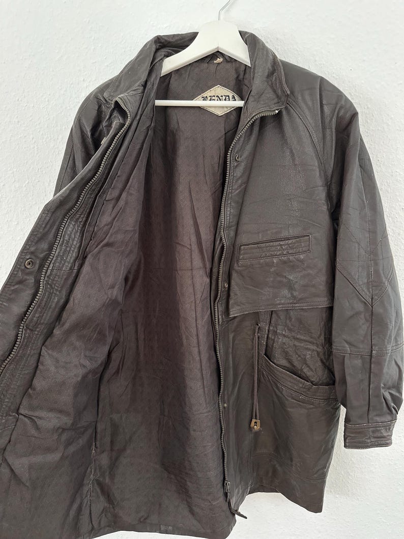 Puede incluir: Chaqueta de cuero marr&oacute;n oscuro con cierre de cremallera y m&uacute;ltiples bolsillos. La chaqueta est&aacute; colgada de una percha blanca. El forro interior es de un tono marr&oacute;n m&aacute;s oscuro. La chaqueta tiene la marca "DENNA" en una etiqueta.