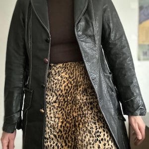 May include: A black leather coat with white stitching and a brown turtleneck. The coat is buttoned and worn over a leopard print skirt. The coat has a classic, tailored design.