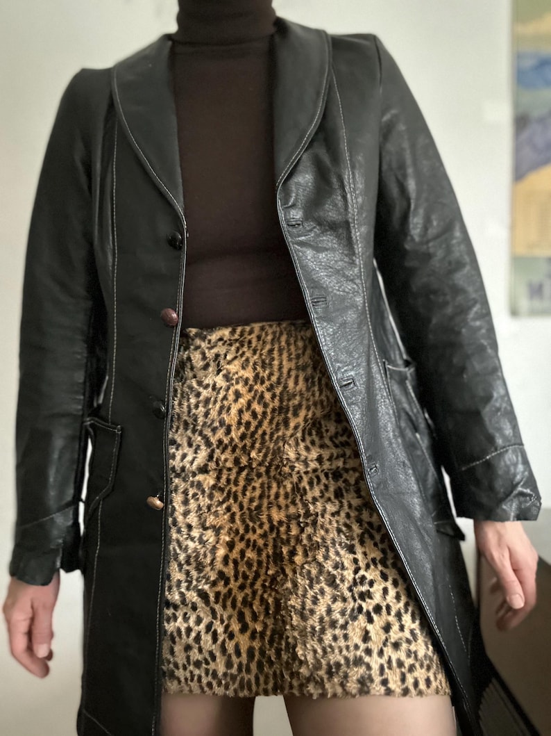 May include: A black leather coat with white stitching and a brown turtleneck. The coat is buttoned and worn over a leopard print skirt. The coat has a classic, tailored design.