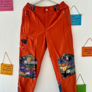 Könnte beinhalten: Leuchtend orange Hose mit bunten Patchwork-Details und Reißverschlusstaschen. Die Hose hängt an einem weißen Kleiderbügel. Sie hat einen Knopfverschluss und elastische Bündchen.