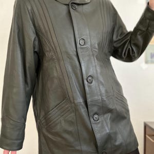 May include: Dark olive green leather jacket with a button-up front, notched collar, and vertical stitched details. The jacket has two front pockets and a tailored fit, suitable for outerwear.