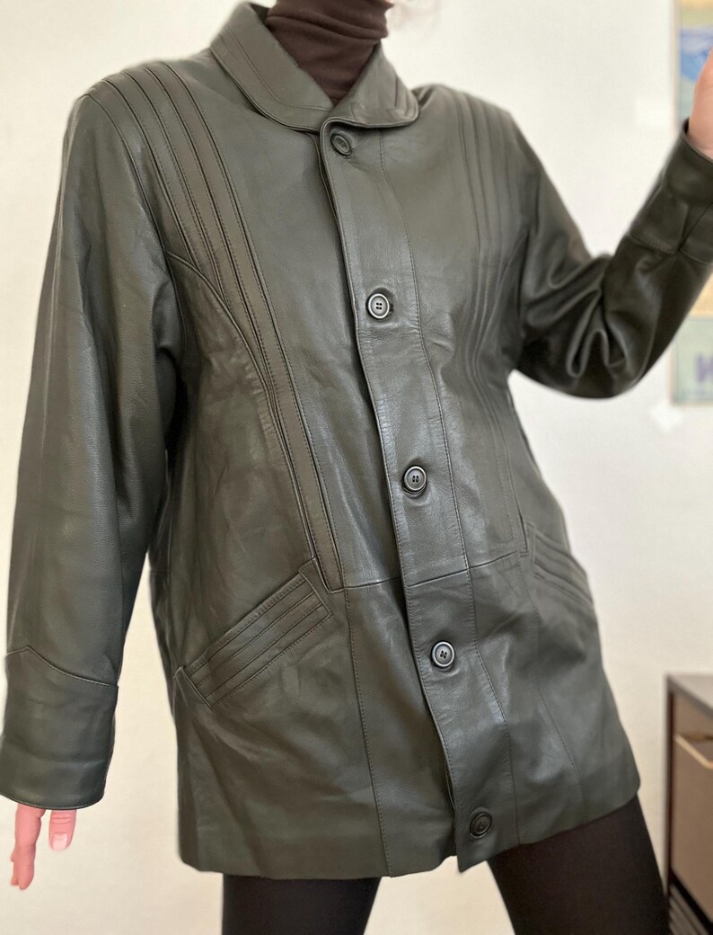 May include: Dark olive green leather jacket with a button-up front, notched collar, and vertical stitched details. The jacket has two front pockets and a tailored fit, suitable for outerwear.
