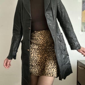 May include: A black leather coat with white stitching and a button closure. The coat is paired with a brown turtleneck and a leopard print skirt. The coat has pockets and a unique asymmetrical hemline.