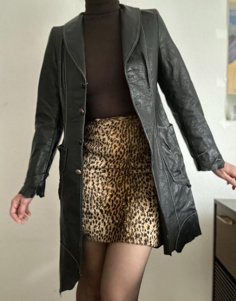 May include: A black leather coat with white stitching and a button closure. The coat is paired with a brown turtleneck and a leopard print skirt. The coat has pockets and a unique asymmetrical hemline.