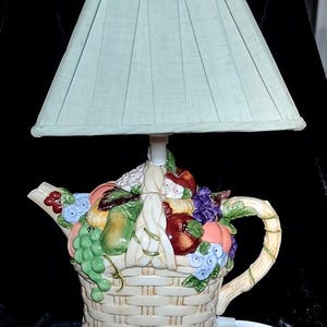 Può includere: Una lampada decorativa a forma di teiera in ceramica con paralume verde chiaro. La base della teiera è a forma di cestino intrecciato traboccante di colorati frutti e fiori in ceramica. Il manico e il beccuccio sono visibili e un cavo bianco si estende dalla base.