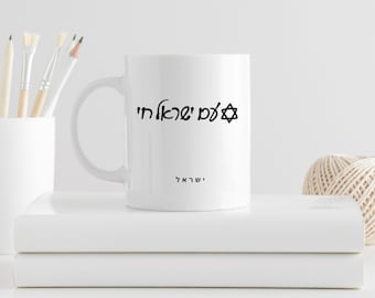 Puede incluir: Taza de cerámica blanca con texto hebreo negro y un diseño de Estrella de David. La taza también tiene la palabra "Israel" impresa. La taza está sobre una pila de libros blancos, con materiales de arte en el fondo.