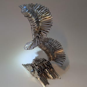 May include: An abstract sculpture made from multiple silver keys. The keys are arranged in a layered, fanned-out design, creating a textured effect. The artwork is set against a neutral background, highlighting the metallic sheen.
