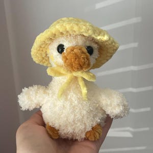 May include: A small, cream-coloured crocheted duckling plush toy featuring a textured, fuzzy yarn body. The duckling wears a bright yellow crocheted sun hat with a matching yellow tie under its chin. It has dark, round eyes and an orange beak.