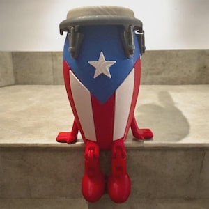 May include: A red, white, and blue conga drum figurine, featuring the flag of Puerto Rico. The drum has a blue top with a white star, red and white stripes, and red legs and feet. The drum is sitting on a step.