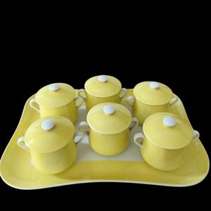 Può includere: Un set di cinque ciotole in ceramica gialla con coperchi, manici bianchi e pomelli bianchi, disposte su un vassoio da portata rettangolare giallo abbinato con angoli arrotondati. Ogni ciotola presenta una finitura liscia e uniforme.