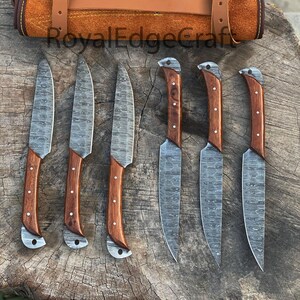 May include: A set of six Damascus steel knives with wooden handles, displayed with a brown leather knife roll. The blades have a unique patterned design. The knife roll is secured with a leather strap and buckle. The text "RoyalEdgeCraft" is visible.