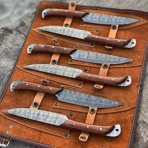 May include: A set of six knives with patterned blades and wooden handles, displayed in a brown leather roll. The blades have a unique textured pattern, and the handles are a dark brown with silver rivets. The leather roll has a buckle closure.