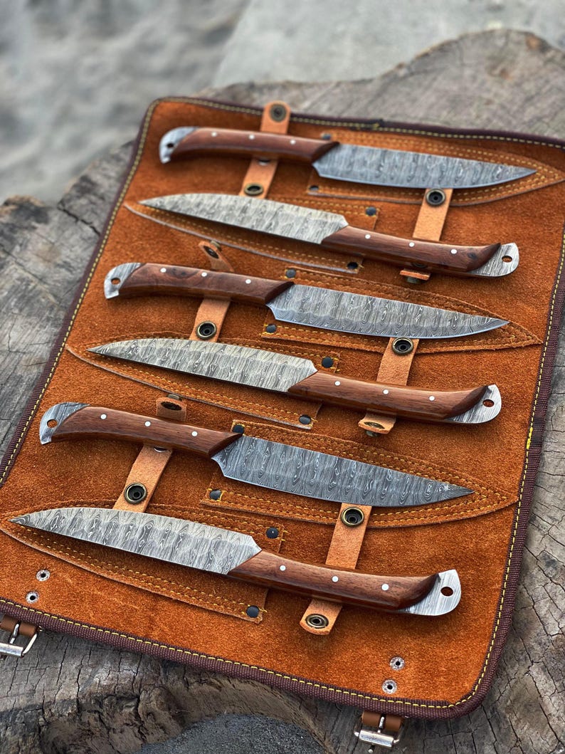 May include: A set of six knives with patterned blades and wooden handles, displayed in a brown leather roll. The blades have a unique textured pattern, and the handles are a dark brown with silver rivets. The leather roll has a buckle closure.