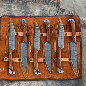 May include: A set of six knives with patterned blades and wooden handles, displayed in a brown leather roll. The blades have a textured appearance, and the handles are a warm brown color. The leather roll has the text "Royal Edge Craft".