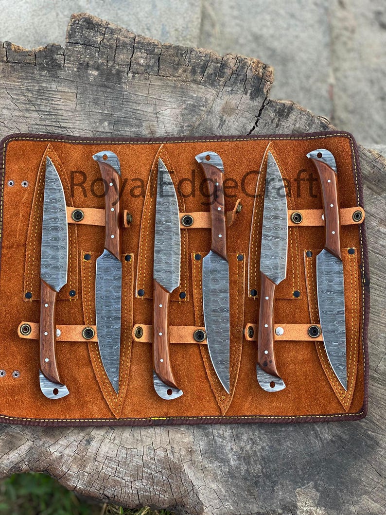 May include: A set of six knives with patterned blades and wooden handles, displayed in a brown leather roll. The blades have a textured appearance, and the handles are a warm brown color. The leather roll has the text "Royal Edge Craft".