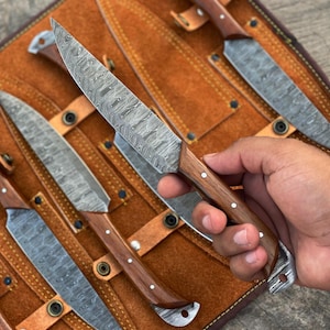 May include: Set of Damascus steel knives with wooden handles and leather sheaths. The blades have a unique patterned design. The knives are displayed in a brown leather roll.