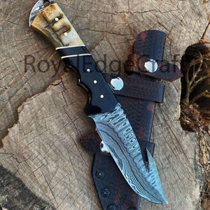 May include: A Damascus steel hunting knife with a black handle and a bone-colored grip. The knife is shown with a brown leather sheath. The blade has a unique pattern. The text "Royal Edge Craft" is visible.