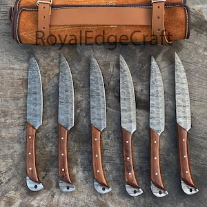 May include: A set of six steak knives with patterned blades and wooden handles, arranged on a wooden surface. The handles are a dark brown color with silver rivets. A brown leather knife roll with buckle closures is at the top. The text "RoyalEdgeCraft" is visible.